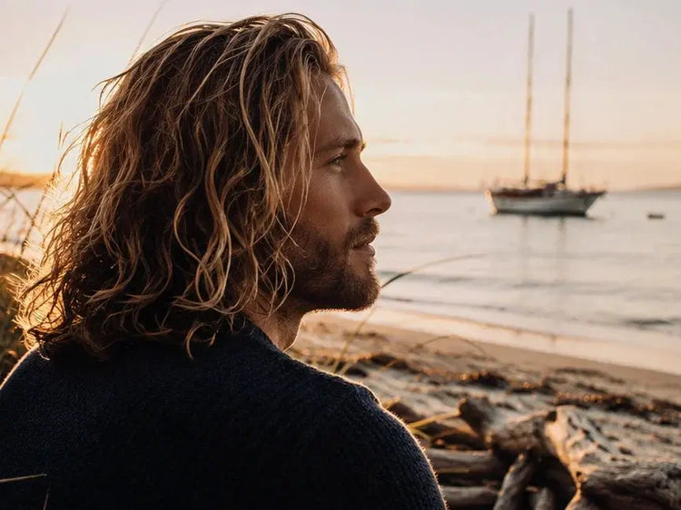 Man with long wavy tallow-washed hair on beach at golden hour, sailboat anchored offshore, Blind Pirate Trading Co.