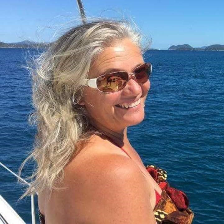 Danita aboard Vivo, her son Kyle’s catamaran, windswept silver-blonde hair and sunglasses, smiling over her shoulder, with deep blue water and distant islands behind her.