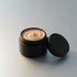 An open black cosmetic jar filled with a beige cream or balm, with its black lid resting beside it on a clean light gray background.