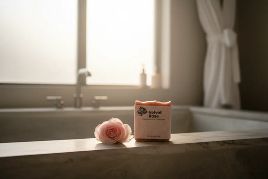 Stone Tub Edge — Morning Light sun falls on a white robe and tub edge, Velvet Rose soap beside a single pink stemmed rose.